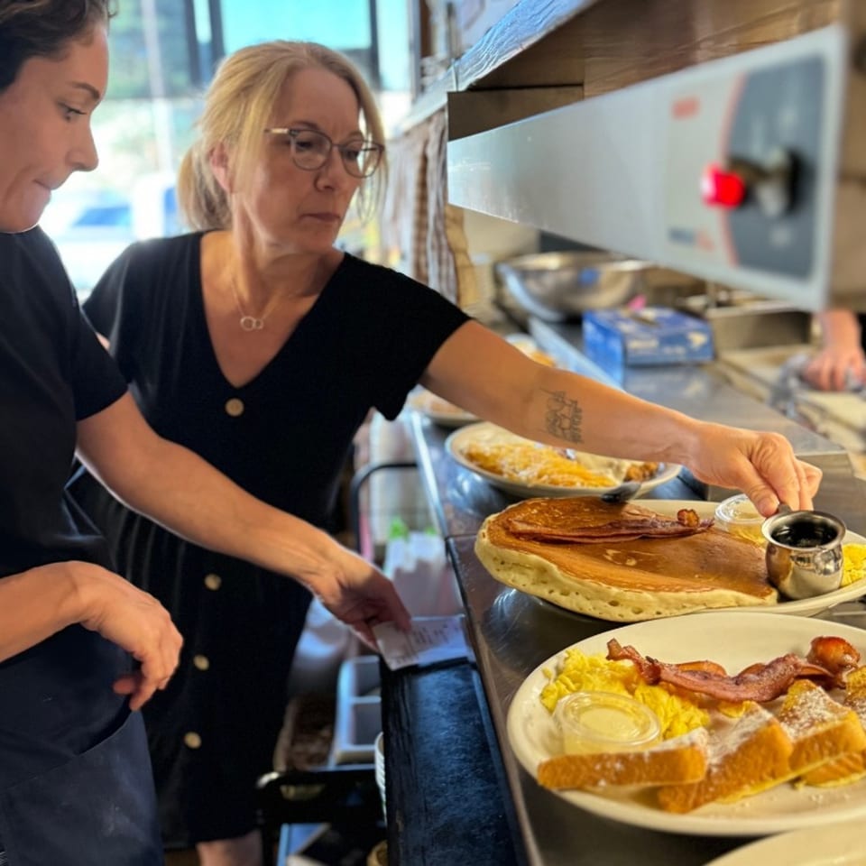 Two women preparing American breakfast plates with pancakes, French toast, eggs, and bacon at Mr T's Family Cafe.