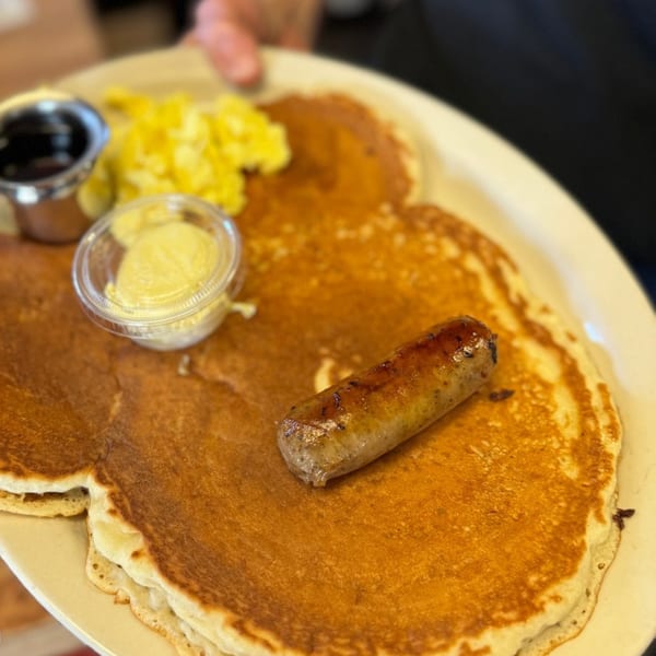 A breakfast plate with three pancakes, a sausage link, scrambled eggs, butter, and syrup.