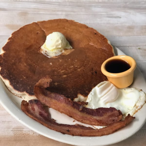 A large pancake with butter, two fried eggs, bacon, and a side of syrup on a white plate.