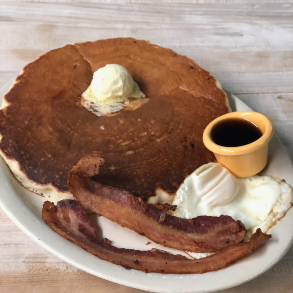 A large pancake with butter, two fried eggs, bacon, and a side of syrup on a white plate.