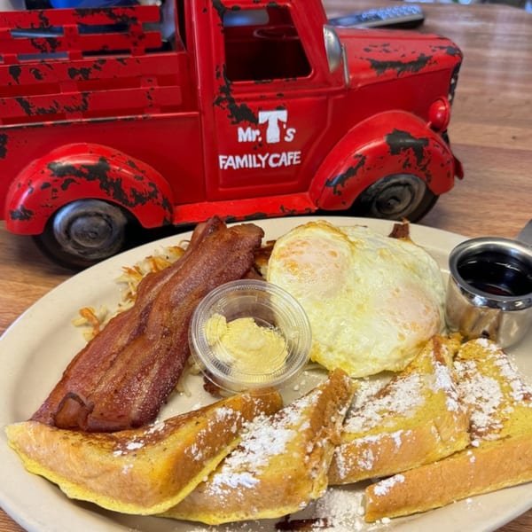 A breakfast plate with French toast, bacon, fried eggs, hash browns, butter, and syrup, with a red toy truck in the background.